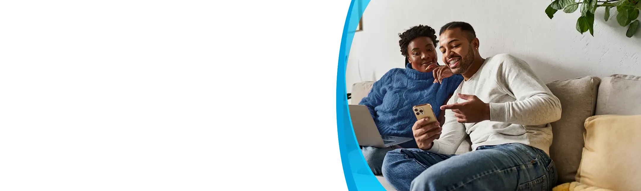 A young couple sits together on a sofa, looking at a smartphone while a laptop rests on one person's lap.